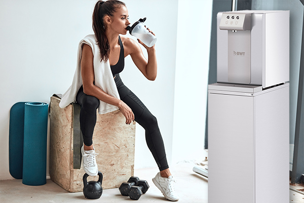 Eine Frau trinkt Wasser in einem Fitnessstudio, neben ihr steht der AQA drink 60 CAS von BWT