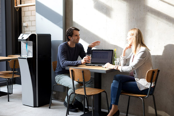 Zwei Personen sitzen auf Stühlen an einem Tisch mit einem Laptop, im Hintergrund ein leerer Stuhl und ein Trinkwasserspender. Die Szene spielt sich in einer hellen Büroumgebung ab.