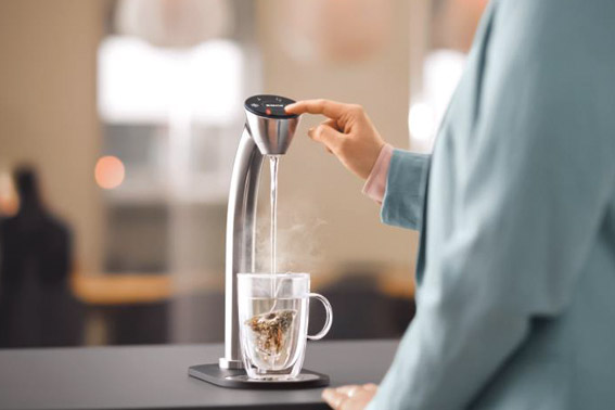 Person bedient einen modernen BRITA Wasserspender, aus dem heißes Wasser in ein Glas mit Teeblättern fließt. Der Wasserspender hat ein schlankes Design und befindet sich auf einer Theke.