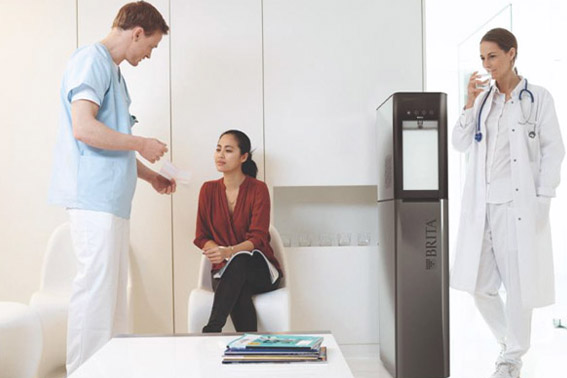 Ein Arzt spricht mit einer Patientin, während eine Ärztin an einem Wasserspender Brita steht und Wasser trinkt.