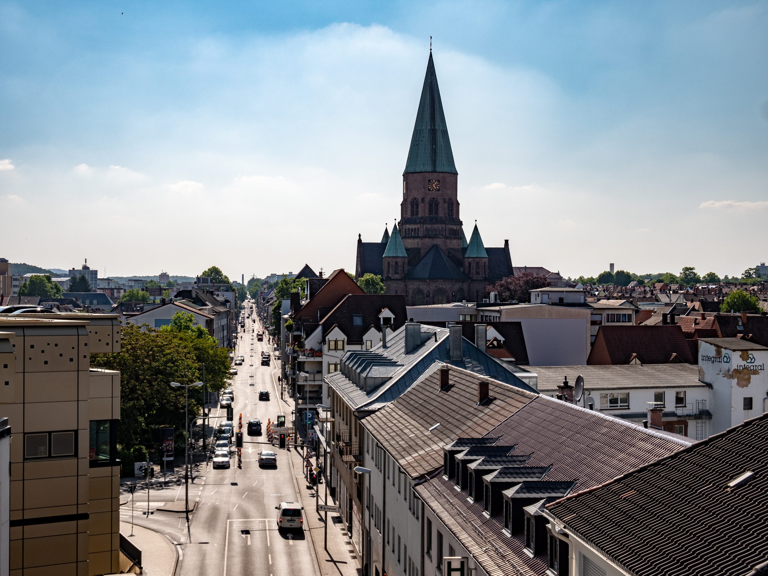 Wasserspender in Kaiserslautern, Trippstadt, Landstuhl, Ramstein-Miesenbach kaufen oder mieten Das Bild zeigt eine städtische Szene in Kaiserslautern, mit der Apostelkirche im Zentrum. Eine lange Straße mit Autos führt an Häusern vorbei und der Himmel ist blau mit leichten Wolken.