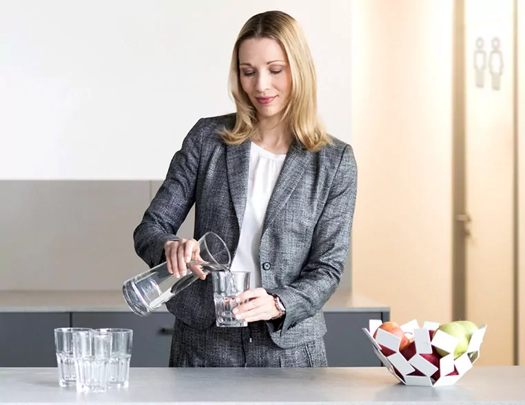 Frau füllt ein Wasserglas mit dem Wasser aus einer Glaskaraffe. Neben ihr steht eine Schale mit frischem Obst.