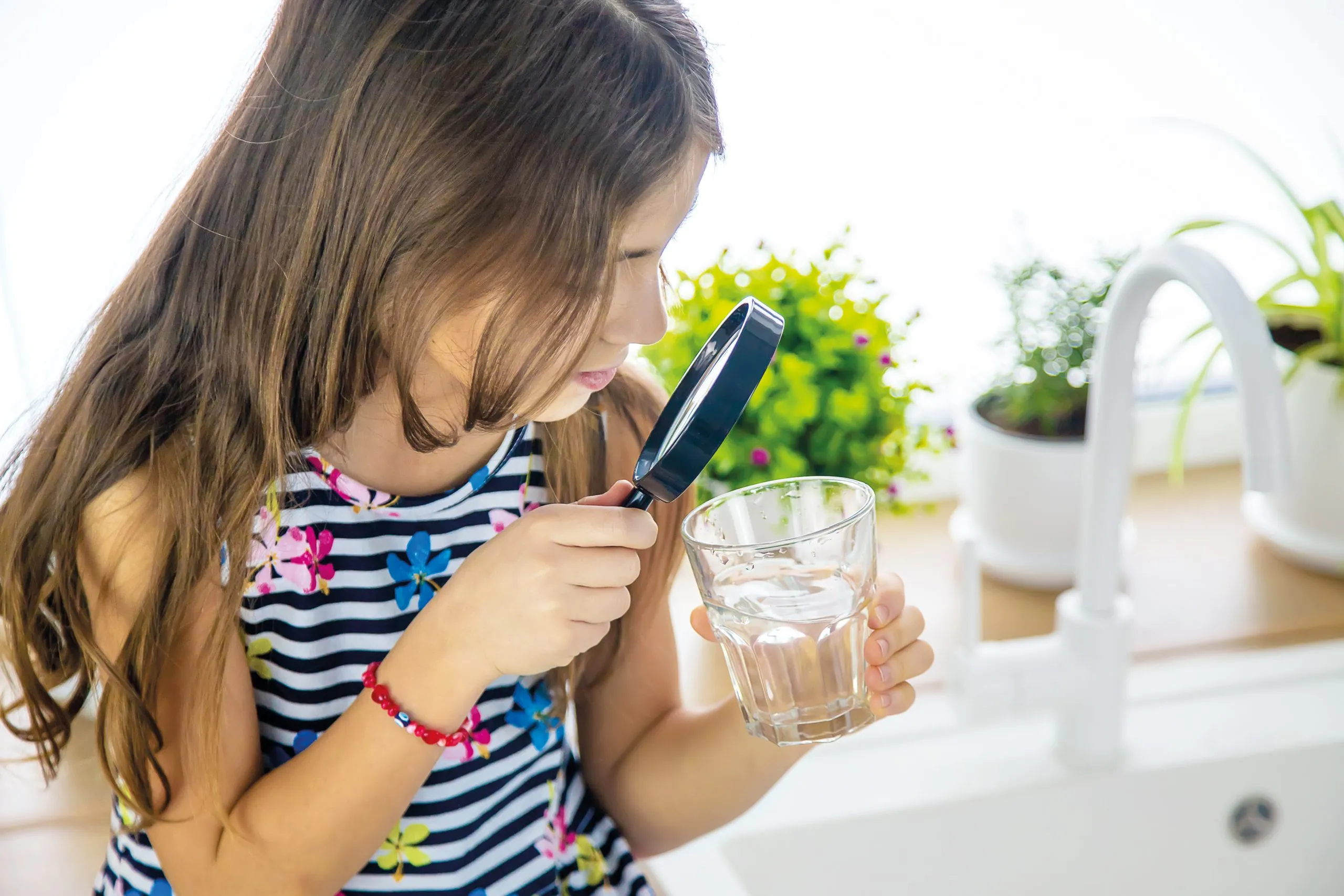 Mädchen, mit langen braunen Haaren, schaut mit einer Lupe in ein mit Wasser gefülltes Wasserglas.