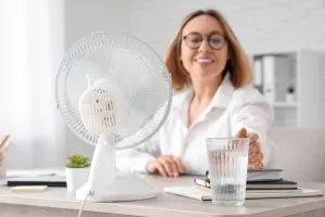Eine Frau greift nach einem kalten Wasserglas. Neben ihr steht ein Ventilator.