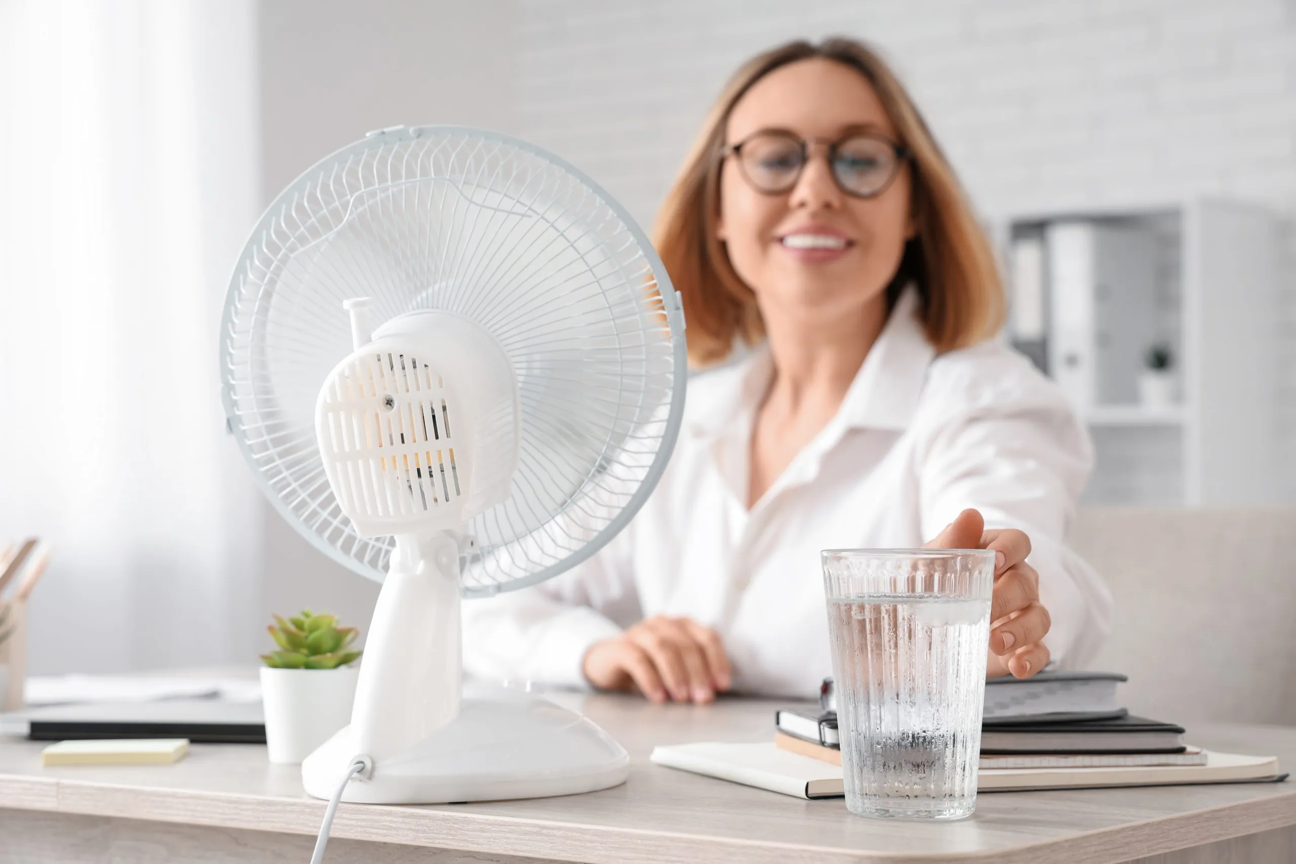 Eine Frau greift nach einem kalten Wasserglas. Neben ihr steht ein Ventilator.
