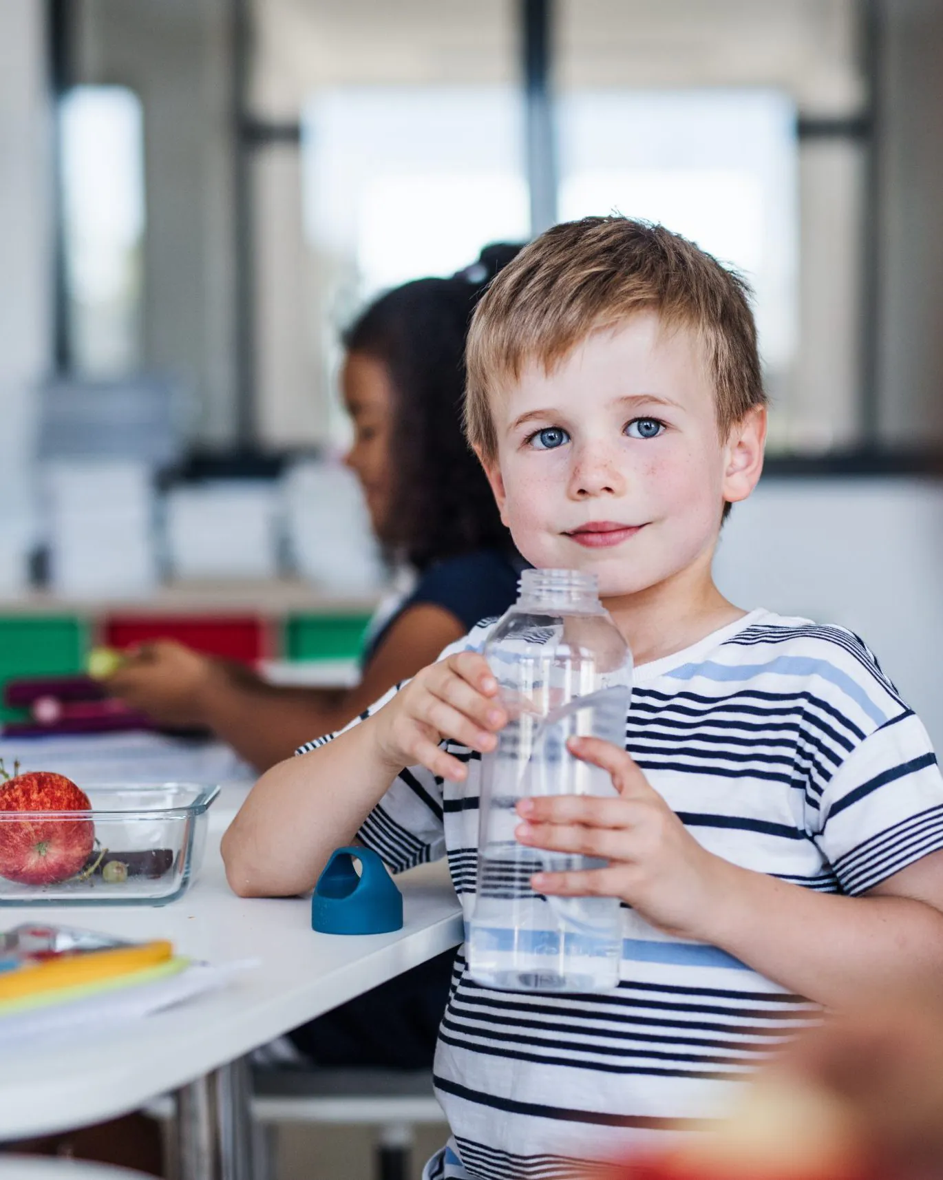 Small school boy sitting at the desk in classroom, drinking water.
